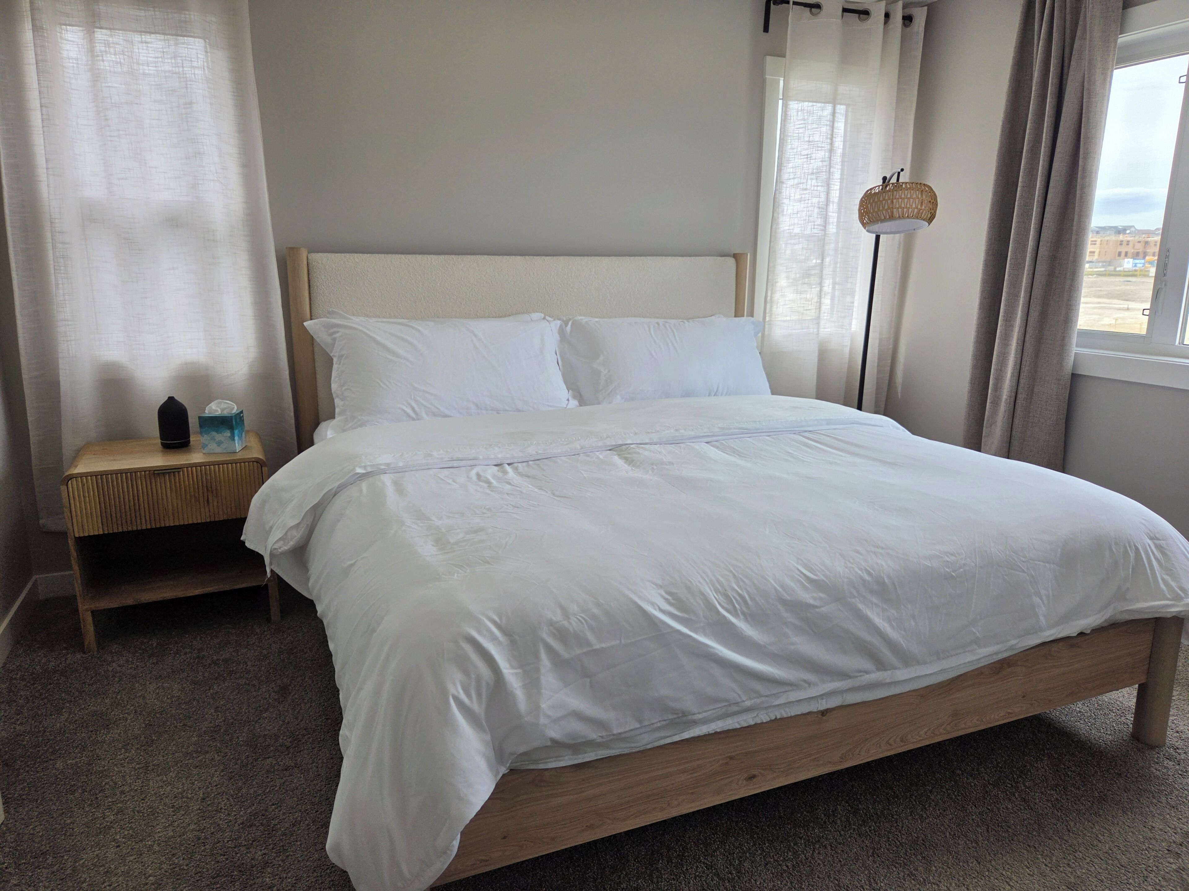 A neatly made bed with white linens, a wooden headboard, and a bedside table with a tissue box and a bottle.