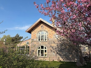 A stone building with a gabled roof and arched windows, surrounded by greenery and flowering trees.