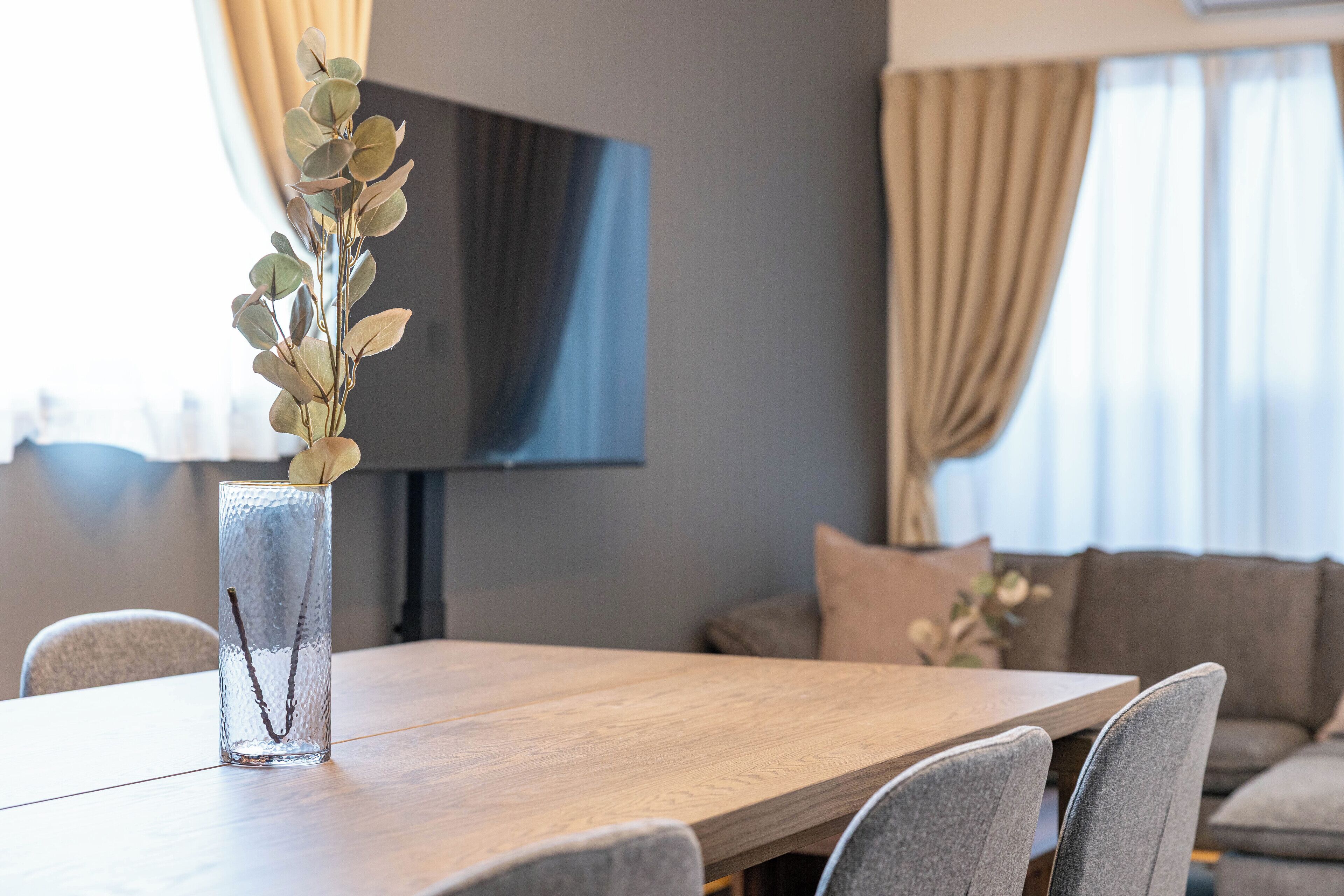 Una mesa de comedor con un florero transparente que contiene una planta, un televisor al fondo y un sofá en primer plano.