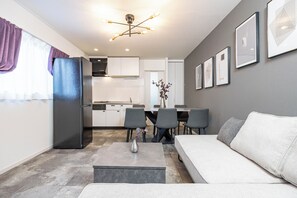 Un salon moderne avec un canapé gris, une table à manger avec des chaises, un coin cuisine avec un réfrigérateur et un luminaire au plafond.