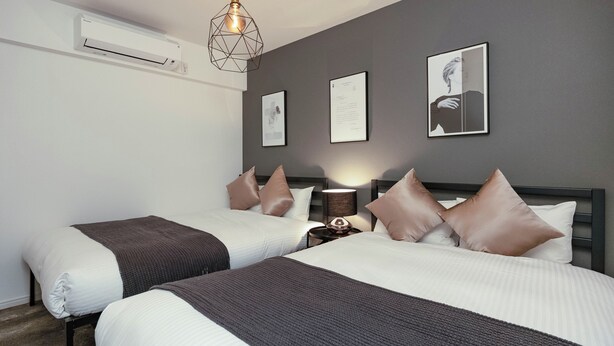A hotel room with two beds, a wall-mounted air conditioner, framed artwork, and a bedside lamp. - Farandole Sanno (Osaka)