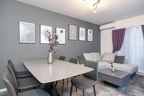 Un salon moderne avec un canapé gris, une table à manger et des chaises, ainsi qu’une table basse. Des tableaux encadrés ornent le mur et un vase rempli de fleurs est posé sur la table.