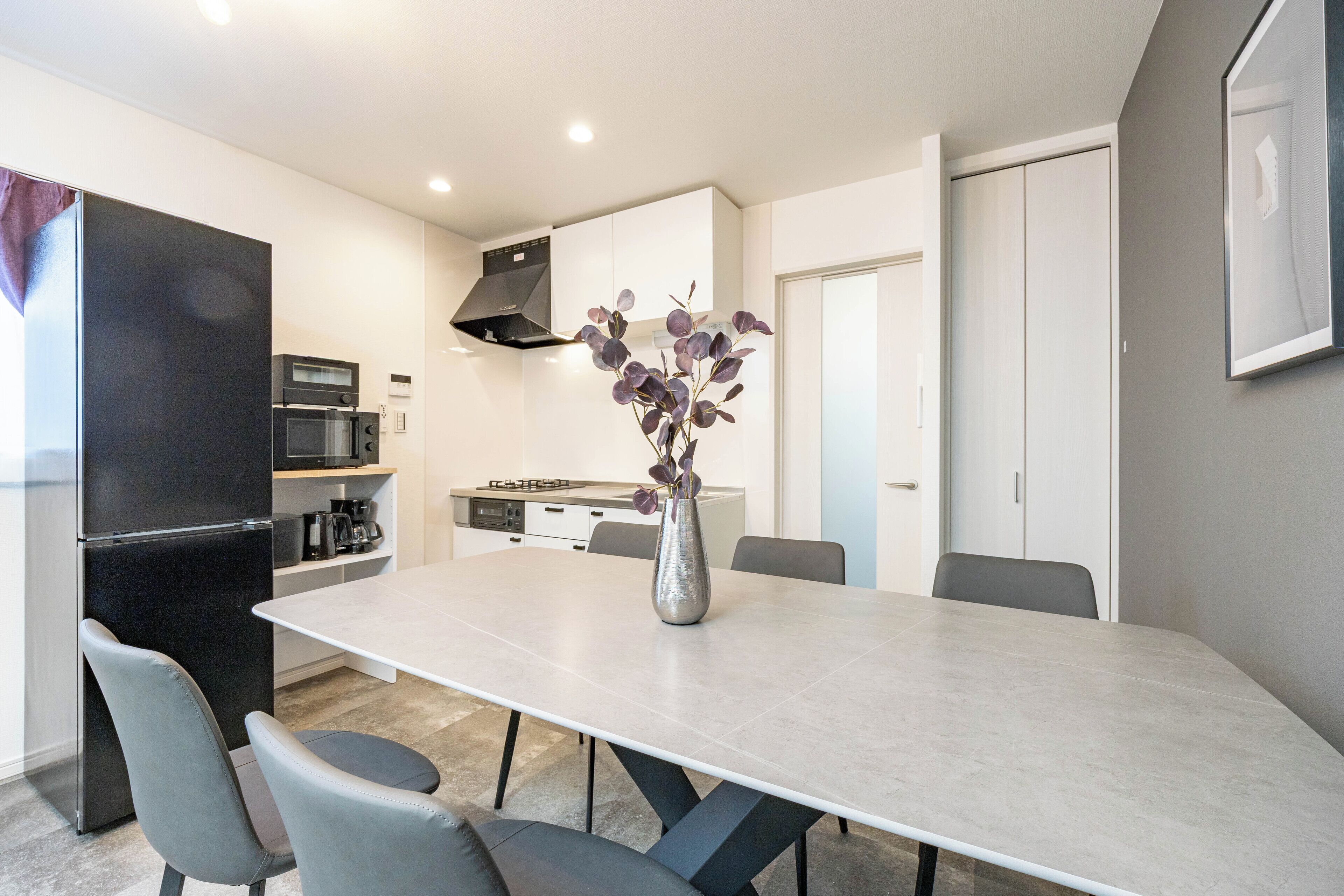 A modern kitchen with a dining table, chairs, a refrigerator, microwave, and a vase with flowers.
