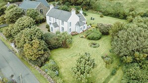 Une grande maison blanche, coiffée d’un toit gris, est entourée d’un jardin bien entretenu et d’arbres.
