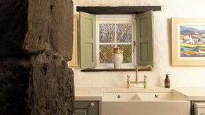 Une cuisine rustique avec un mur en pierre, une fenêtre avec des volets verts, un évier blanc et un tableau encadré accroché au mur.