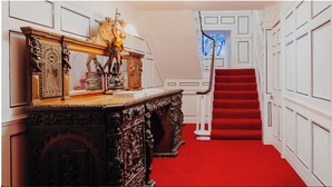 Un couloir recouvert d’une moquette rouge menant à un escalier, agrémenté de meubles en bois richement décorés et doté de murs aux panneaux blancs.