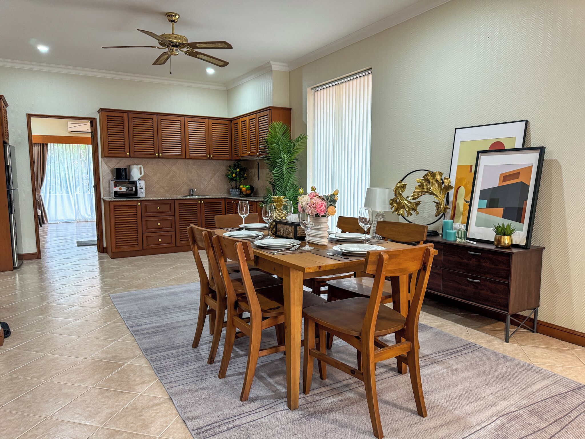 Un comedor con mesa y sillas de madera, un ventilador de techo y una cocina al fondo.