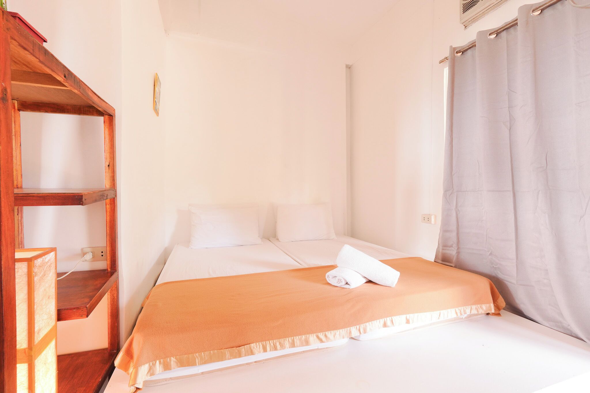 A single bed with a white mattress and orange bedding, a wooden shelf unit, and a grey curtain.