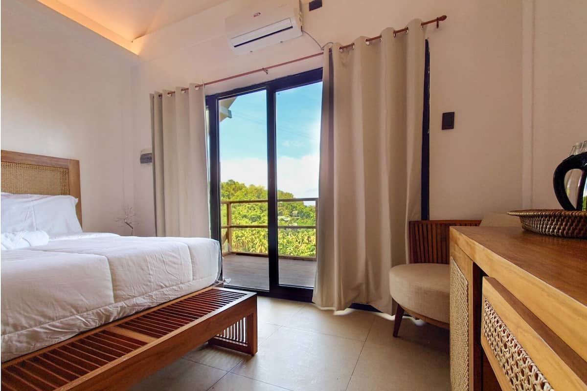 Ein Schlafzimmer mit Bett, einer Bank, einem Sessel, einem Schreibtisch und Blick auf Grün hinter einer Schiebetür.