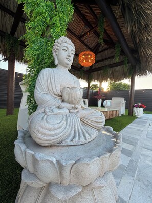 Eine stehende Buddha-Statue auf einem Sockel, im Hintergrund ein strohgedecktes Dach und eine Hängeleuchte.