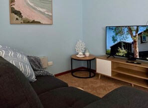 A living room with a sofa, a small round table with a decorative item, a television displaying a house, and a bookshelf. - Howrah Apartment (Howrah)