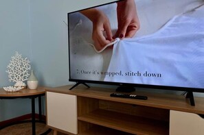 A television displaying a tutorial on sewing, with a wooden TV stand and a small table with decorative items. - Howrah Apartment (Howrah)