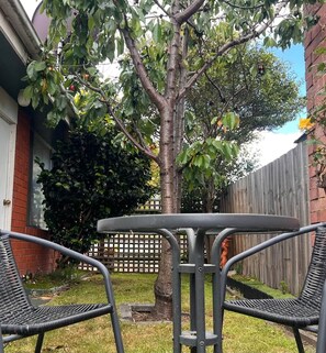 Un patio con una mesa metálica y dos sillas, rodeado por un muro de ladrillo y una valla de madera.