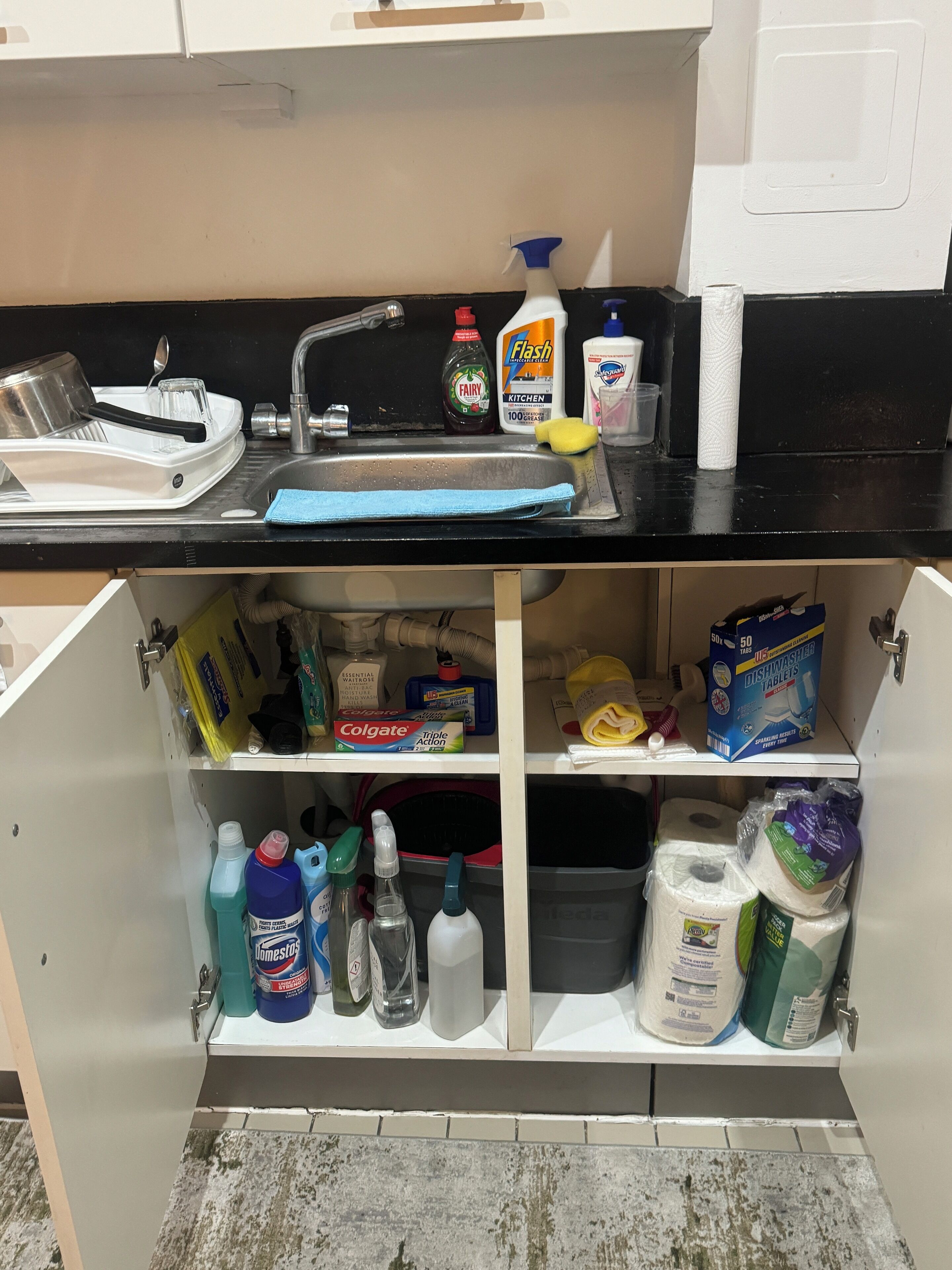 Een keuken met een spoelbak, afwassysteem en diverse schoonmaakmiddelen.