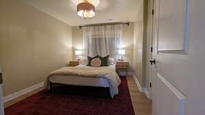 Ein Schlafzimmer mit einem Bett, zwei Nachttischen, einem Fenster mit durchsichtigen Vorhängen und einer Deckenleuchte.