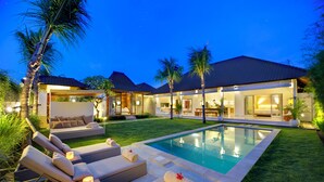 A poolside area with lounge chairs, a pool, and a building with large windows and a covered patio. - Sahana Villas (Seminyak)