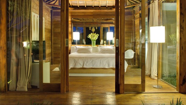 A bedroom with a large bed, a nightstand, a lamp, and a window with curtains. - Sahana Villas (Seminyak)