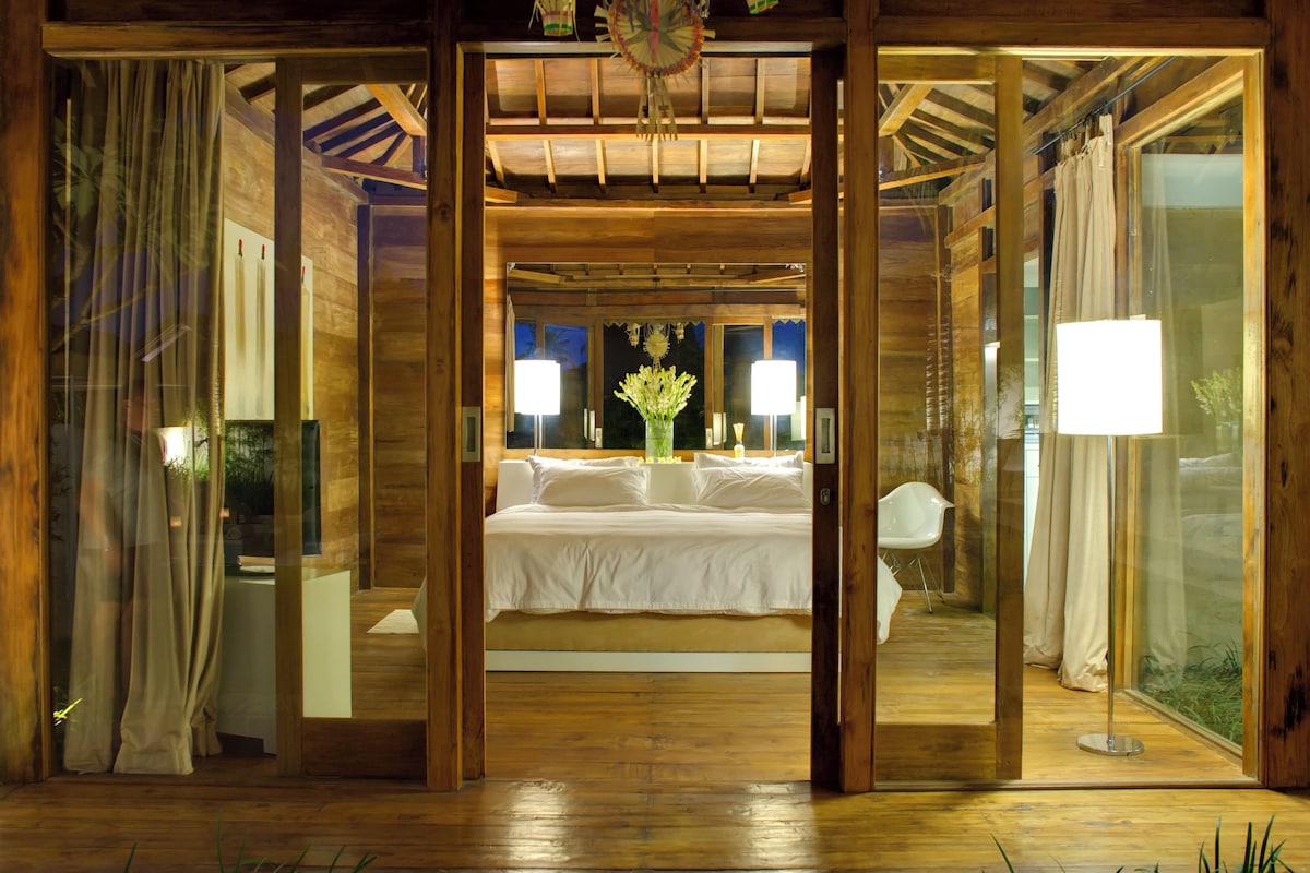 A bedroom with a large bed, a nightstand, a lamp, and a window with curtains.
