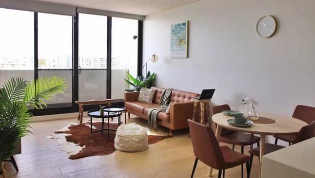 Un salon moderne avec un canapé, une table basse, une table à manger et des chaises. On peut voir un ordinateur portable sur le canapé, une horloge au mur et une plante sur le sol.