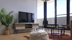 Un salon moderne avec un grand téléviseur à écran plat, un meuble en bois, un fauteuil noir, une petite table ronde et une plante en pot.