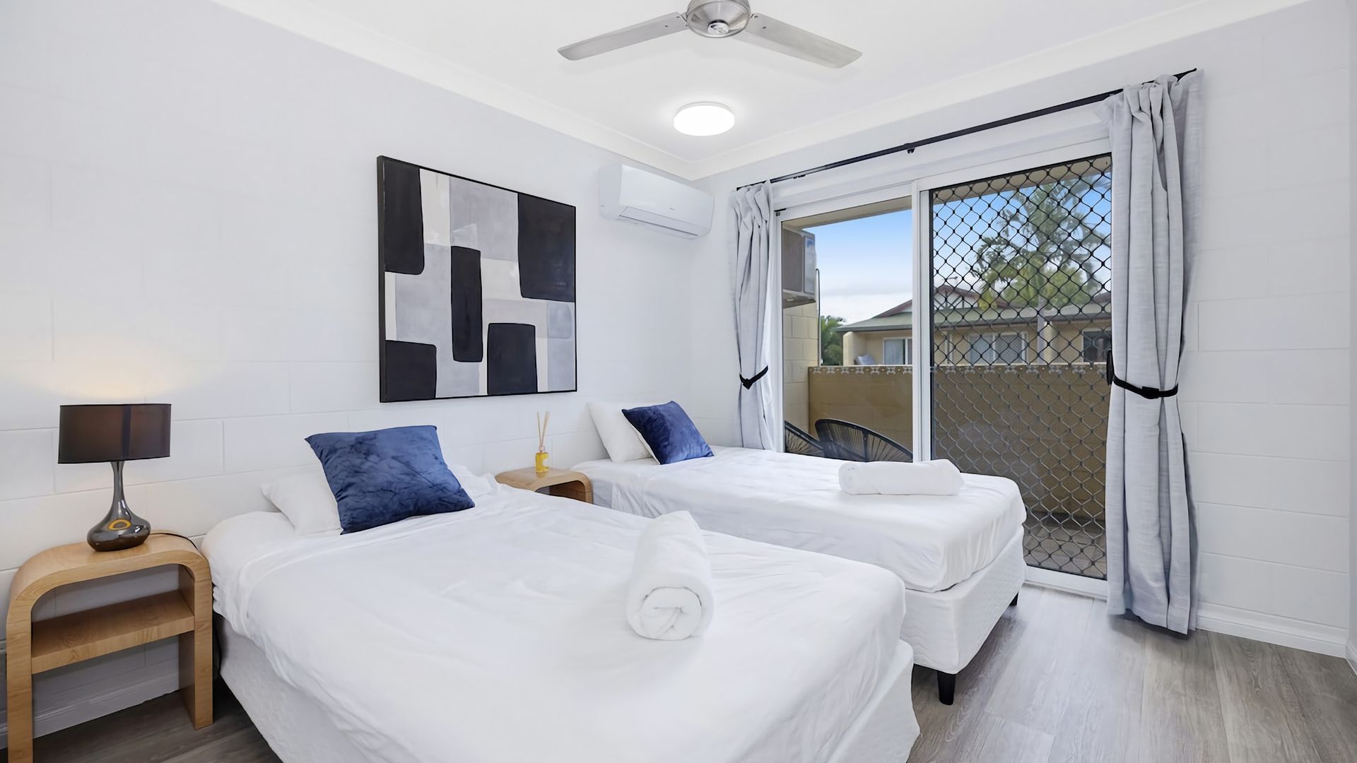 A hotel room with two beds, a ceiling fan, an air conditioning unit, a window with curtains, and a view of the outside.