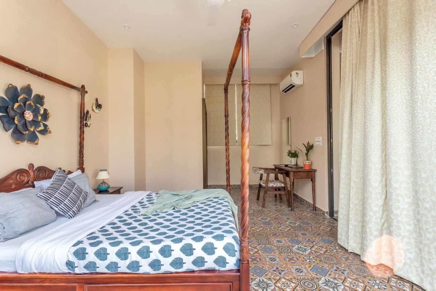 A four-poster bed with a patterned bedspread, a desk with a chair, and a large decorative wall piece.
