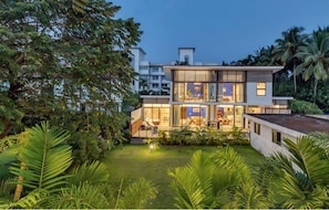 Une maison moderne à deux étages, dotée de grandes fenêtres, d’une pelouse bien entretenue et entourée d’une végétation tropicale luxuriante.