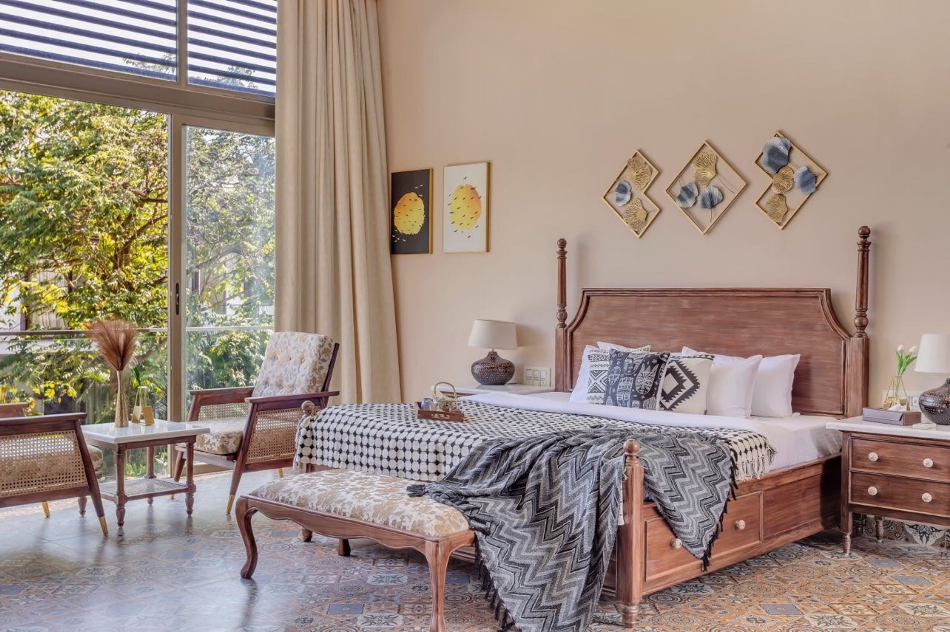 A spacious bedroom with a large bed, a wooden nightstand, a bedside table, and a chair. There are three framed artworks on the wall, and the room has a large window with a view of trees.