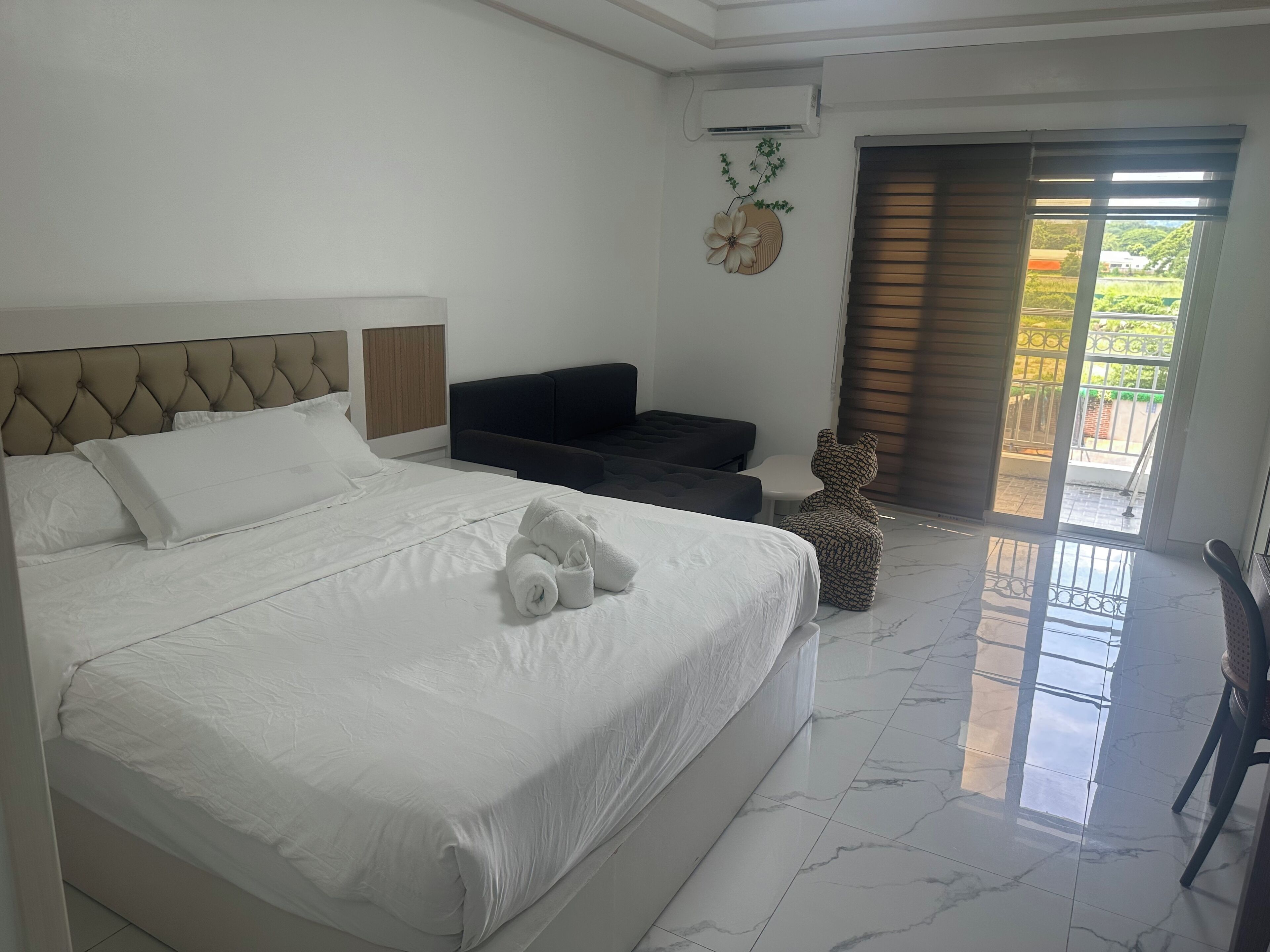 A hotel room with a large bed, a sofa, a small table, a decorative wall piece, and a view of the outdoors through a sliding door.