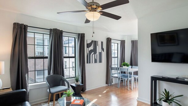 Un salon moderne doté d’un ventilateur de plafond, d’une table basse en verre, d’un canapé noir et d’un coin repas avec une table et des chaises blanches.