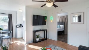 Un salon moderne doté d’un ventilateur de plafond, d’un téléviseur à écran plat, d’une console noire agrémentée d’une plante, d’une machine à café et d’un coin repas avec des chaises.