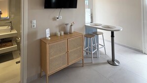 Ein modernes Hotelzimmer mit einem an der Wand befestigten Flachbildfernseher, einem Holztischchen mit Dekorationselementen, einem runden Esstisch mit Hocker und einer Decke mit einem linearen Muster.