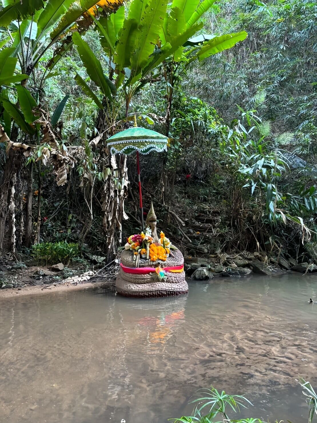 Una estructura tradicional con un paraguas verde, decorada con flores y una estatua, ubicada junto a un río tranquilo.