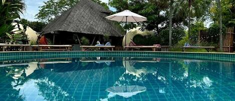 A clear blue pool with a tiled deck, surrounded by palm trees and a thatched-roof hut.