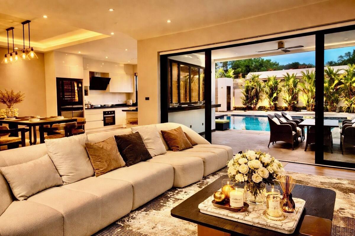 Ein modernes Wohnzimmer mit einer Couch, einem Essbereich und einer Küche. Ein großes Glasfenster führt zu einem Poolbereich mit Gartenmöbeln.