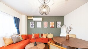 A modern living room with a sectional sofa, a round wooden coffee table, a dining table with chairs, and a hanging light fixture. - Loger Tengachaya (Osaka)