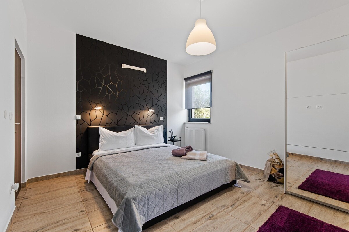 A modern bedroom with a geometric-patterned headboard, a large bed with a quilt, a bedside table, a hanging light fixture, a window, and a mirror.