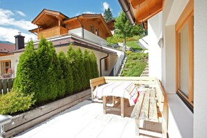 A wooden deck with a table and benches, surrounded by greenery and a building with a sloped roof. - Chalet in Saalbach Near Ski Lift (Saalbach-Hinterglemm)