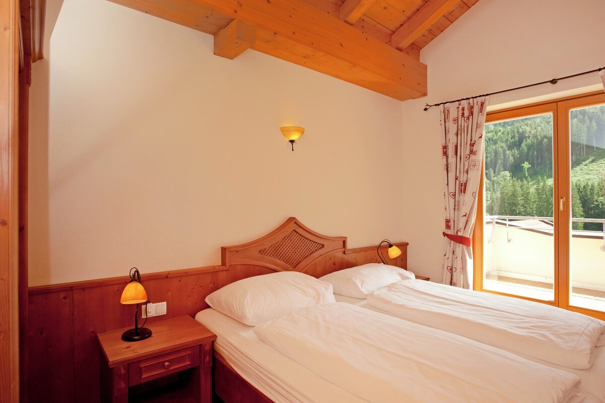 A bedroom with two beds, wooden headboards, a nightstand with a lamp, a wall-mounted light, and a window with curtains.