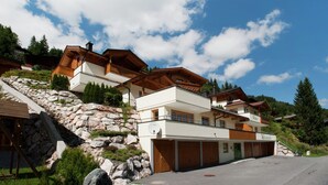 A multi-story chalet with a stone retaining wall, wooden balconies, and a garage. - Chalet in Saalbach Near Ski Lift (Saalbach-Hinterglemm)