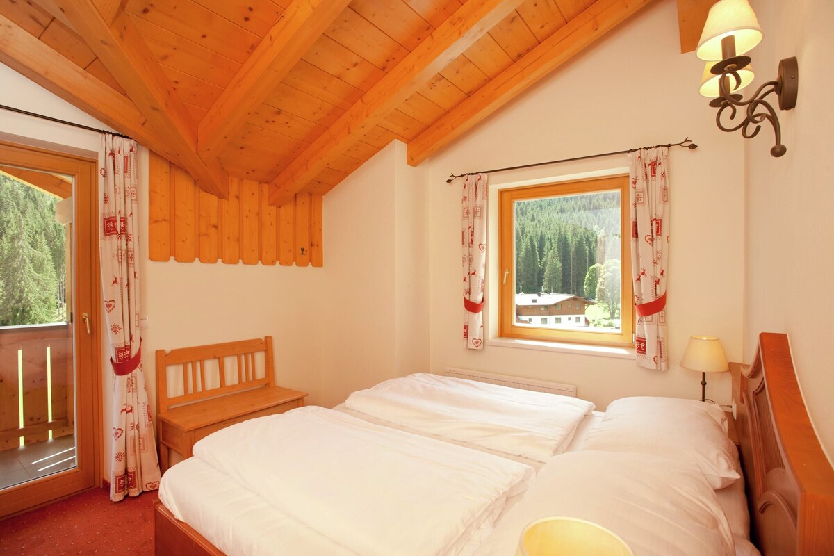A bedroom with a wooden ceiling, a large window with curtains, a bed, and a nightstand with a lamp.