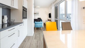 Eine moderne Küche mit weißen Schränken, einer schwarzen Arbeitsplatte und einem eingebauten Ofen. Es gibt eine Essecke mit Tisch und Stühlen, und ein großes Fenster sorgt für natürliches Licht.