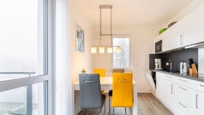 Eine moderne Küche mit weißen Schränken, einem Essbereich mit weißem Tisch und zwei Stühlen, einer gelb und einer grau, und einem großen Fenster, das für natürliches Licht sorgt.