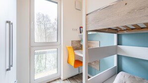 Ein modernes, kleines Zimmer mit einem Etagenbett, einem Fenster mit Blick auf Bäume, einem kleinen Schreibtisch mit Stuhl und einem Regal mit Büchern und einer Lampe.