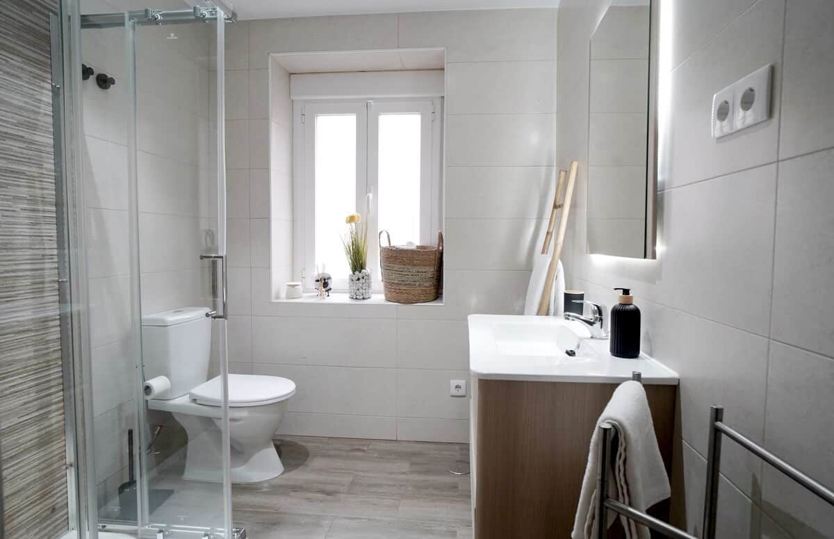 A modern bathroom with a glass shower enclosure, a toilet, a sink with a mirror, and a window with a basket.