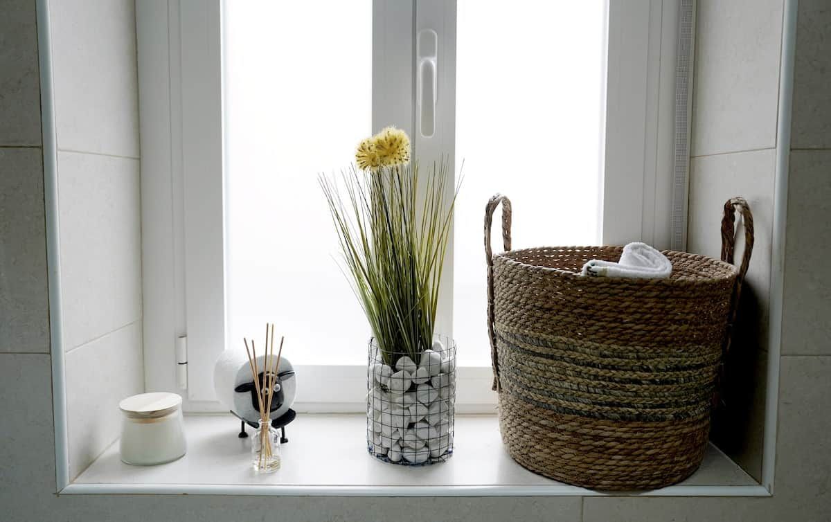 Un lavabo da bagno con una vasca bianca in ceramica, un cesto intrecciato e una pianta decorativa in un cestino metallico.