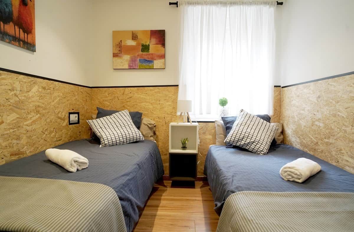 Une chambre avec deux lits, une petite table de chevet avec une lampe, une fenêtre avec des rideaux et un tableau au mur.