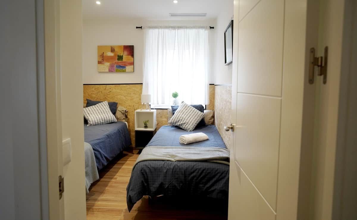 Ein Schlafzimmer mit zwei Betten, einem Nachttisch mit Lampe, einem Fenster mit Vorhängen und einem Gemälde an der Wand.
