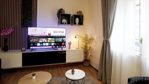 Un salon avec un téléviseur affichant les mots « OLIVIA » et « Enfants » à l’écran, une petite table avec une plante, une lampe décorative et une plante en pot sur une étagère.