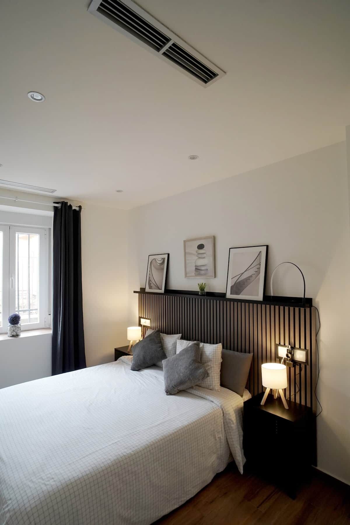 Ein Schlafzimmer mit einem Bett, Nachttischlampen, einem Fenster mit Vorhängen und Wandbildern.